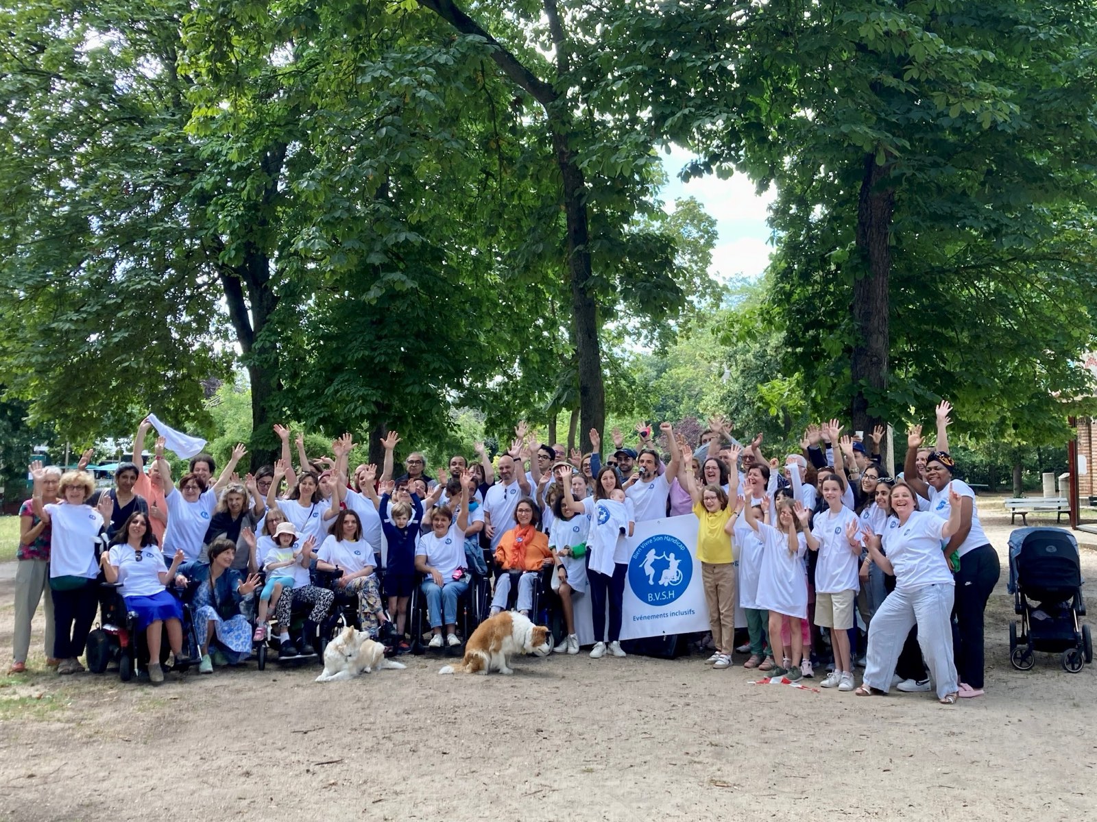 Photo de groupe de l’association Bien Vivre Son Handicap dans un parc : une soixantaine de bénévoles et participants en t-shirts blancs lèvent les bras en souriant autour de la banderole B.V.S.H « Événements inclusifs », avec plusieurs personnes en fauteuil roulant et deux chiens au premier plan.