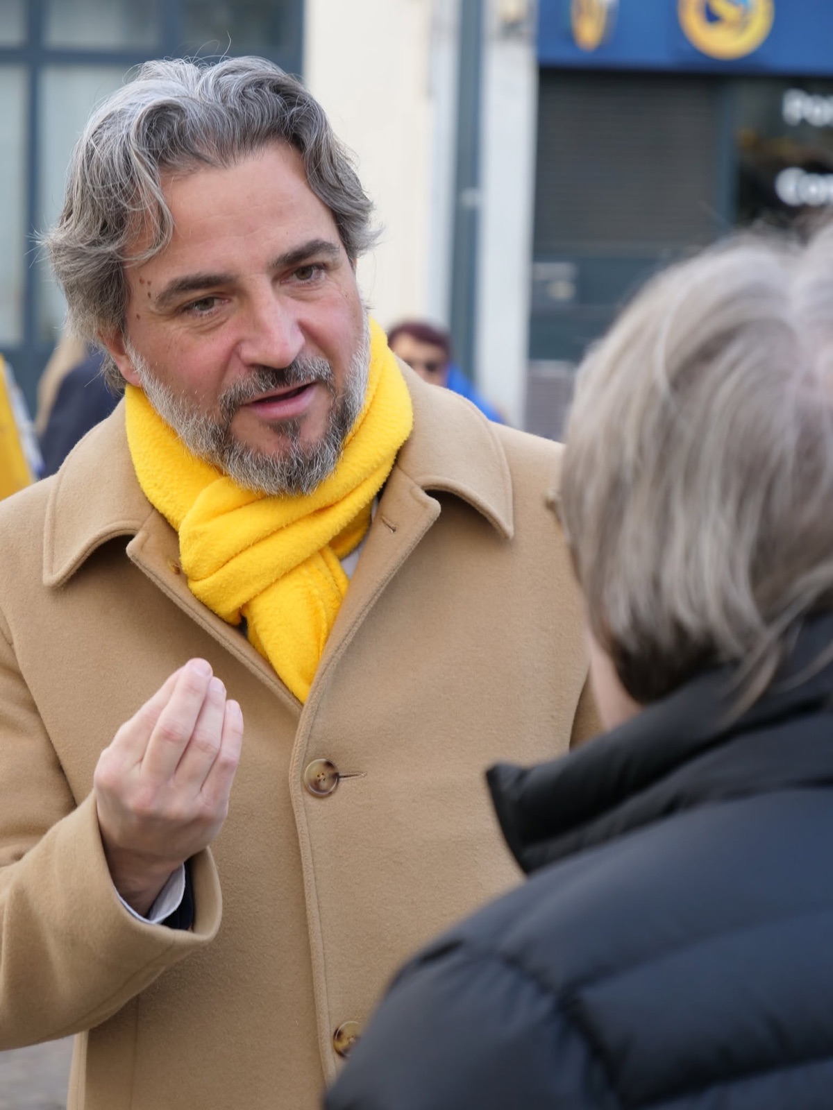 Vincent Julé, en manteau camel et écharpe jaune, en pleine discussion avec une habitante dans la rue pendant la campagne municipale.
